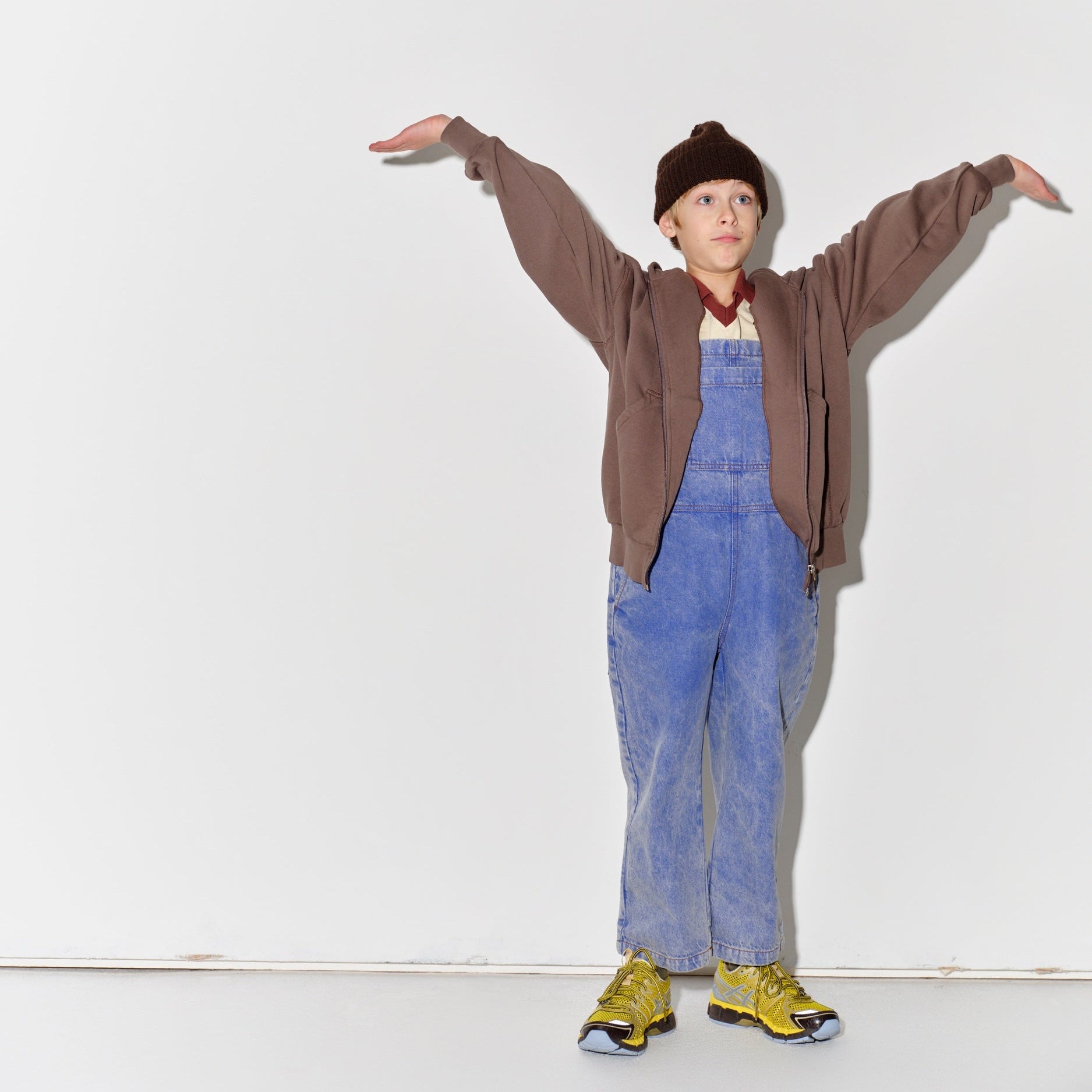 Kid wearing a Main Story dark brown knit beanie, falcon brown hoodie, blue denim overalls, and yellow shoes on a white background