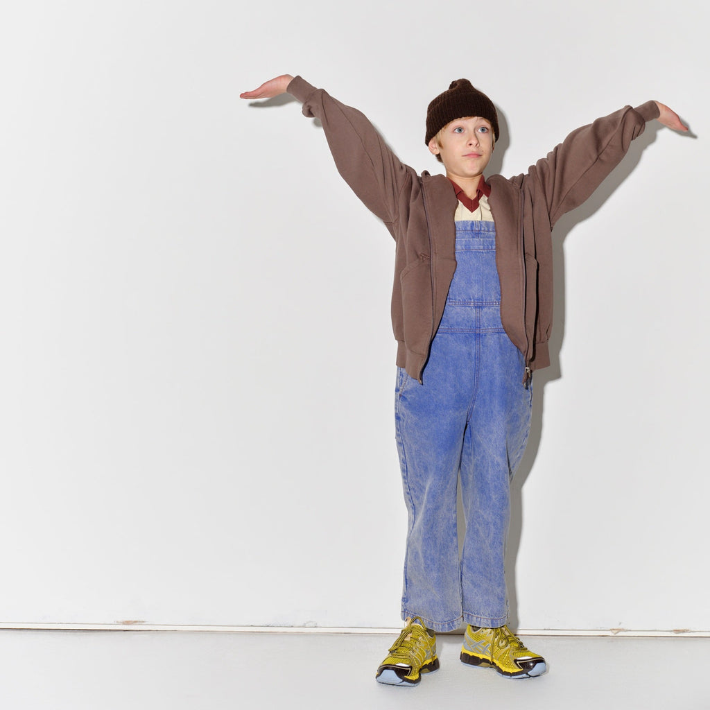 Kid wearing a Main Story dark brown knit beanie, falcon brown hoodie, blue denim overalls, and yellow shoes on a white background
