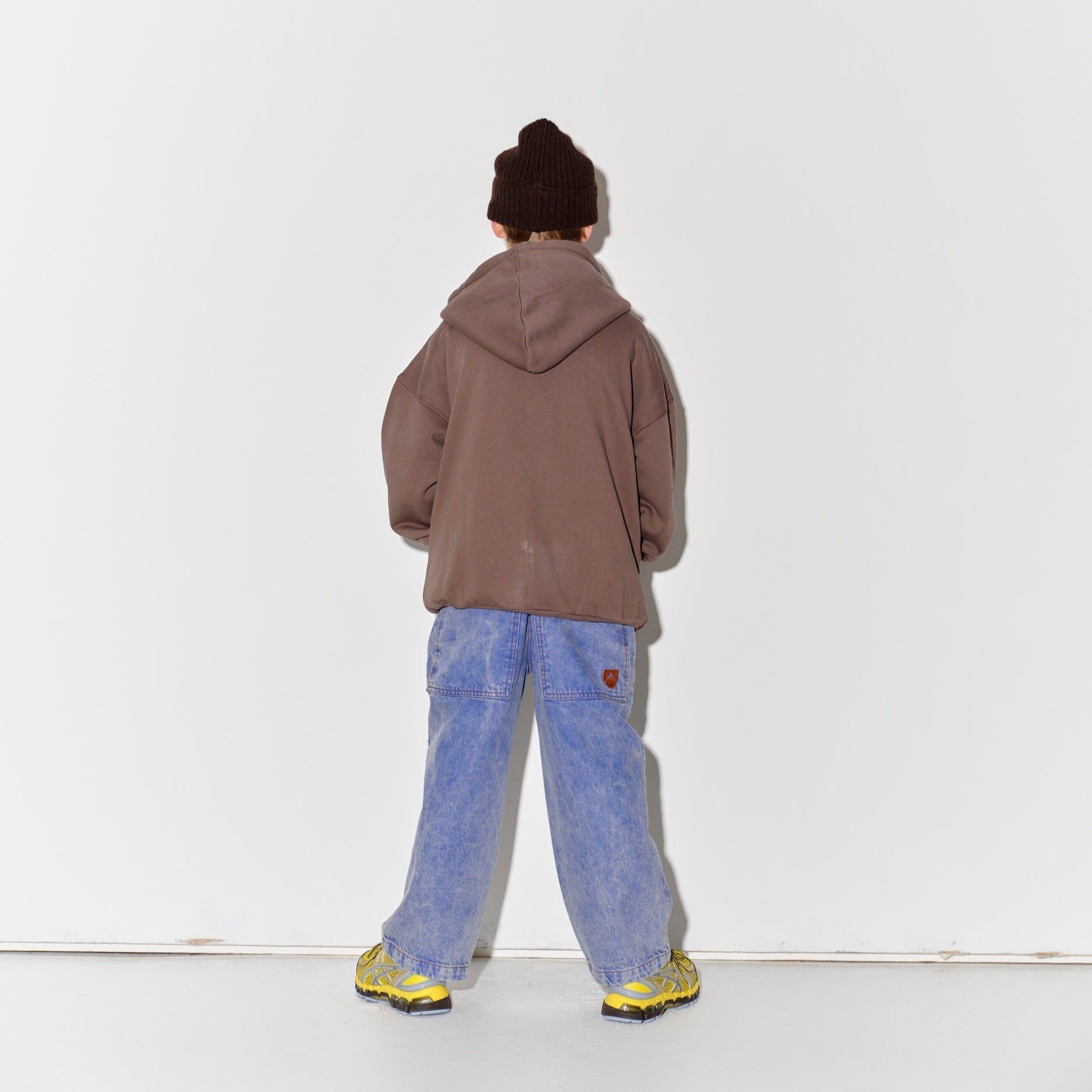 Kid wearing a Main Story dark brown knit beanie, falcon brown hoodie, blue denim overalls, and yellow shoes on a white background