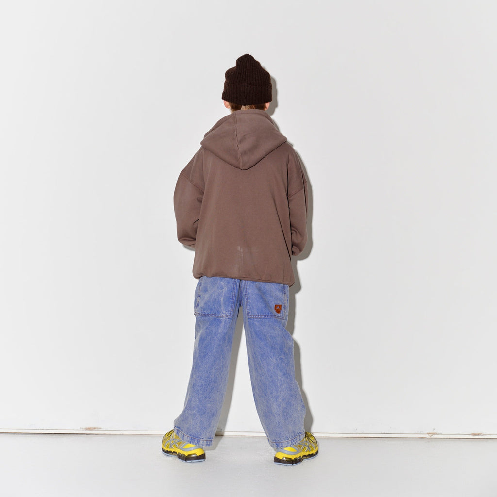Kid wearing a Main Story dark brown knit beanie, falcon brown hoodie, blue denim overalls, and yellow shoes on a white background