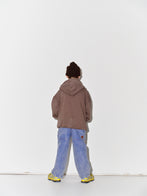 Kid wearing a Main Story dark brown knit beanie, falcon brown zip hoodie sweatshirt, blue denim overalls, and yellow shoes on a white background
