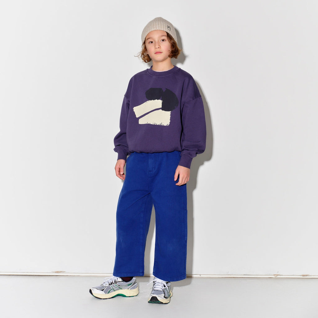 Child wearing a Main Story beige knit beanie, purple sweatshirt with a graphic design, and true blue painter cargo pants