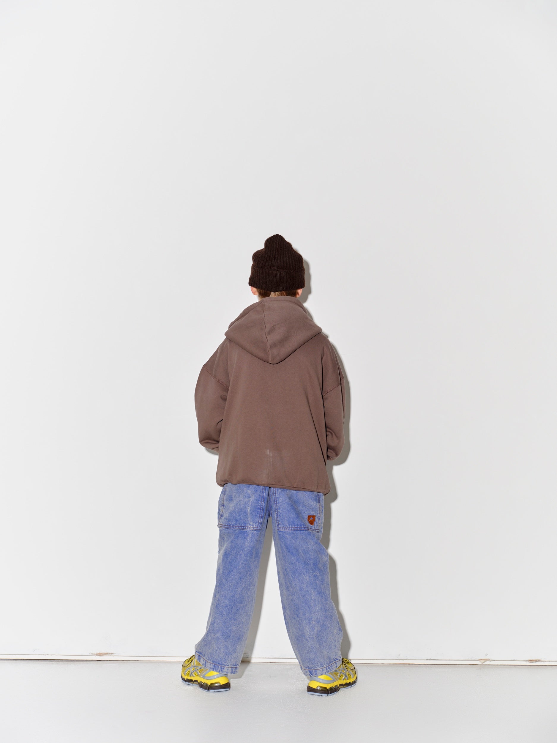 Kid wearing a Main Story dark brown knit beanie, falcon brown zip hoodie sweatshirt, blue denim overalls, and yellow shoes on a white background
